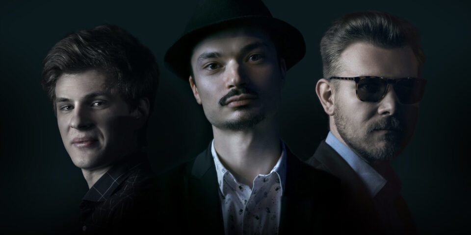 Trzech mężczyzn w ujęciu portretowym na czarnym tle. Pierwszy z nich ma jasne włosy i jest uśmiechnięty, drugi ma mały wąsik i czarny kapelusz, trzeci ma ciemne okulary. Wszyscy mają koszule i marynarki. Tło jest czarne.