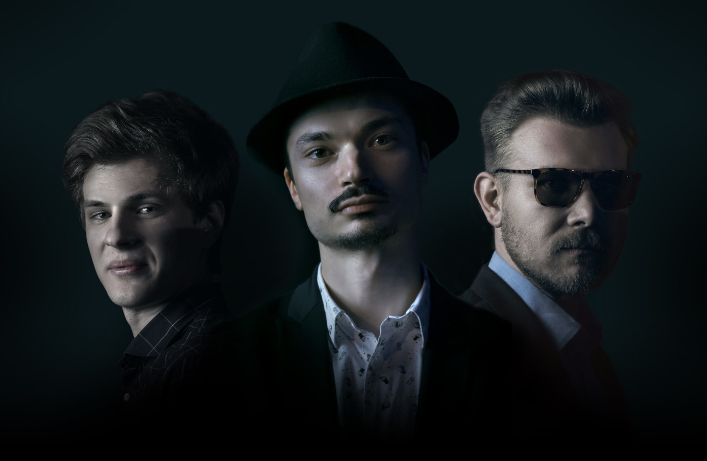 Trzech mężczyzn w ujęciu portretowym na czarnym tle. Pierwszy z nich ma jasne włosy i jest uśmiechnięty, drugi ma mały wąsik i czarny kapelusz, trzeci ma ciemne okulary. Wszyscy mają koszule i marynarki. Tło jest czarne.