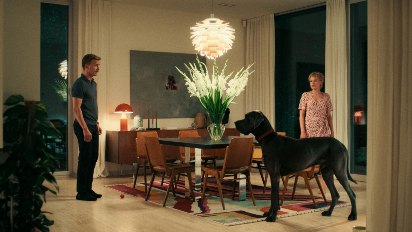 Zdjęcie dwóch osób i psa w dużym salonie. Po lewej stronie stoi mężczyzna , po prawej kobieta. Przed kobietą stoi duży, czarny pies. Na środku jest stół, dookoła niego krzesła. Na stole jest wazon z dużym bukietem białych kwiatów, nad nim ozdobny żyrandol. W tle ściana z dużym obrazem i wysokimi okami i zasłonami.