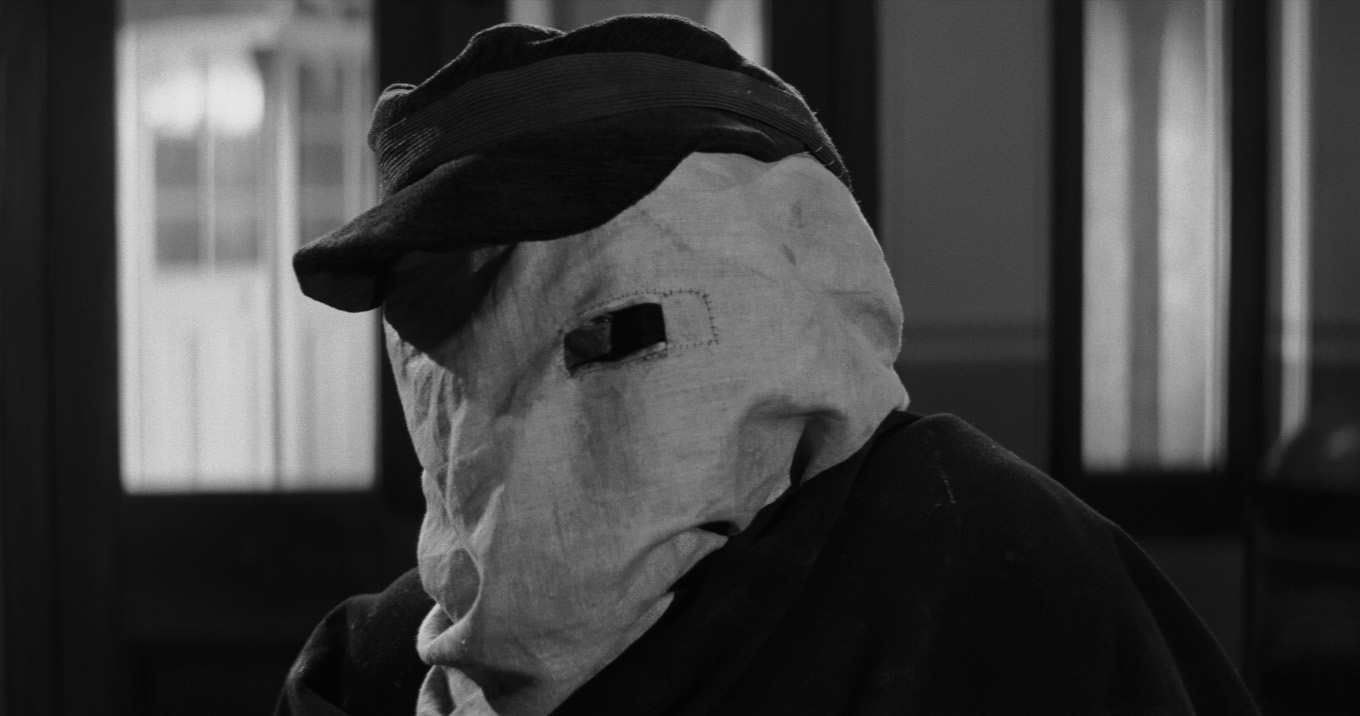 Czarno-białe zdjęcie portretowe postaci z założonym na głowę jasnym, płóciennym workiem. W worku jest wycięta niewielka prostokątna dziura odsłaniająca oko. Postać ma ciemny strój i ciemną czapkę z daszkiem. W tle wnętrze pomieszczenia.