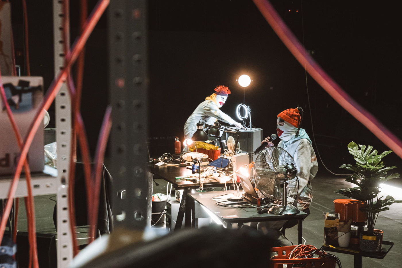 Zdjęcie dwóch osób w zagraconej przestrzeni. Osoby są ubrane w białe kombinezony i czarno-czerwone czapki. Na twarzach mają maski klaunów. Maski są w formie kominiarek. Na scenie są dwa stoły, na których jest mnóstwo różnego rodzaju przedmiotów. Na podłodze też stoją różne rzeczy.