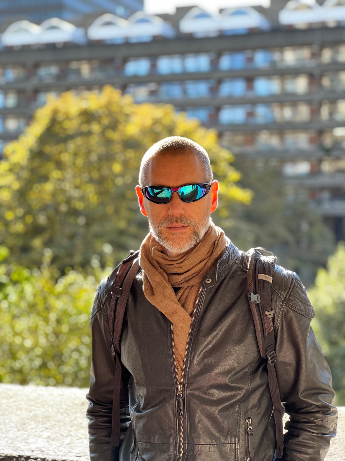Zdjęcie Tomasza Ulanowskiego w ujęciu od pasa w górę. Jest ubrany w brązową, skórzaną kurtkę i szalik w kolorze wielbłądzim. Ma ciemne okulary z niebieskimi refleksami. W rozmytym tele są drzewa i wysoki budynek z dużą ilością okien.