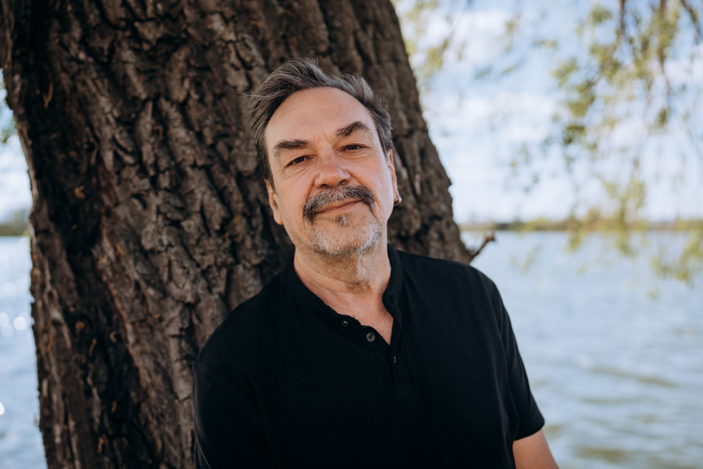 Zdjęcie portretowe uśmiechniętego mężczyzny. Jest ubrany w czarną koszulkę z krótkimi rękawami. Ma siwawe włosy, wąsy i krótki zarost. W tle po lewej bardzo grube drzewo, po prawej krajobraz z jeziorem i niebem.