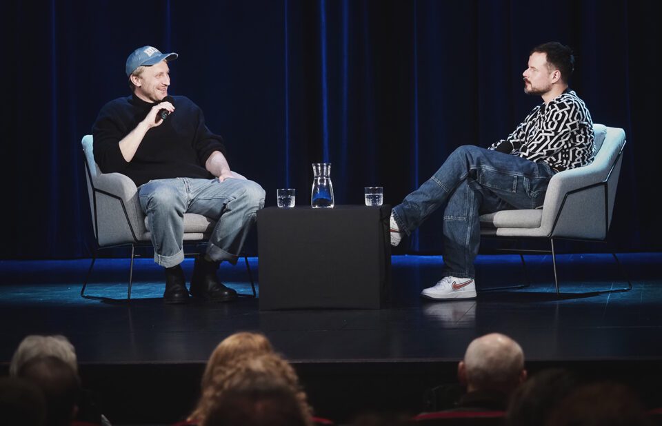 Zdjęcie dwóch mężczyzn siedzących na przeciwko siebie w fotelach na scenie. Ujęcie zza głów widzów z pierwszego rzędu.