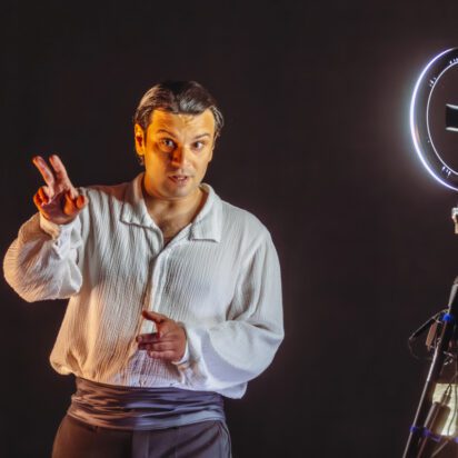 Obraz Główny: Zdjęcie mężczyzny w ujęciu od bioder w górę. Ma ciemne spodnie i jasną koszulę. Mężczyzna wyciąga przed siebie rękę, drugą trzyma blisko ciała na wysokości brzucha. Po prawej stronie stoi lampa na statywie, dająca blade światło. Tło czarne.