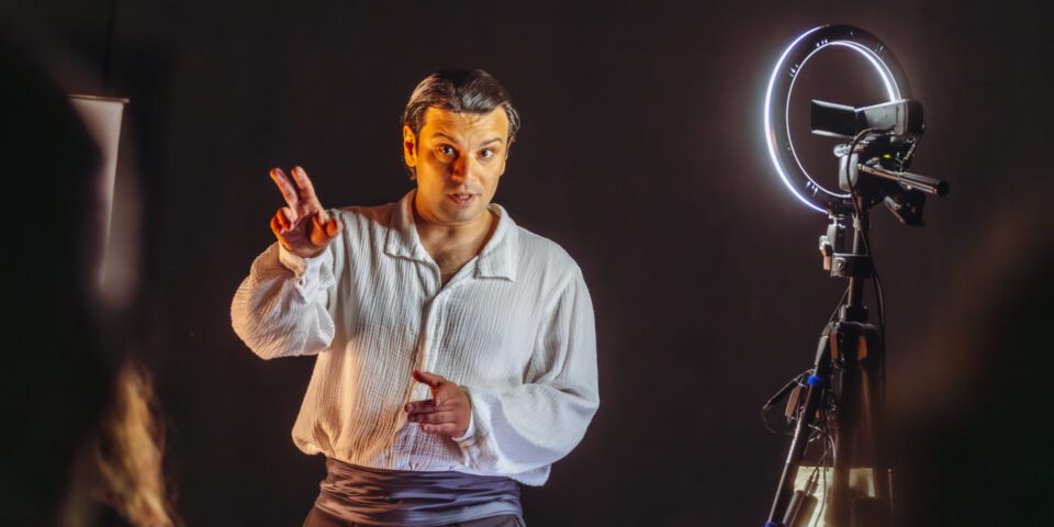 Zdjęcie mężczyzny w ujęciu od bioder w górę. Ma ciemne spodnie i jasną koszulę. Mężczyzna wyciąga przed siebie rękę, drugą trzyma blisko ciała na wysokości brzucha. Po prawej stronie stoi lampa na statywie, dająca blade światło. Tło czarne.