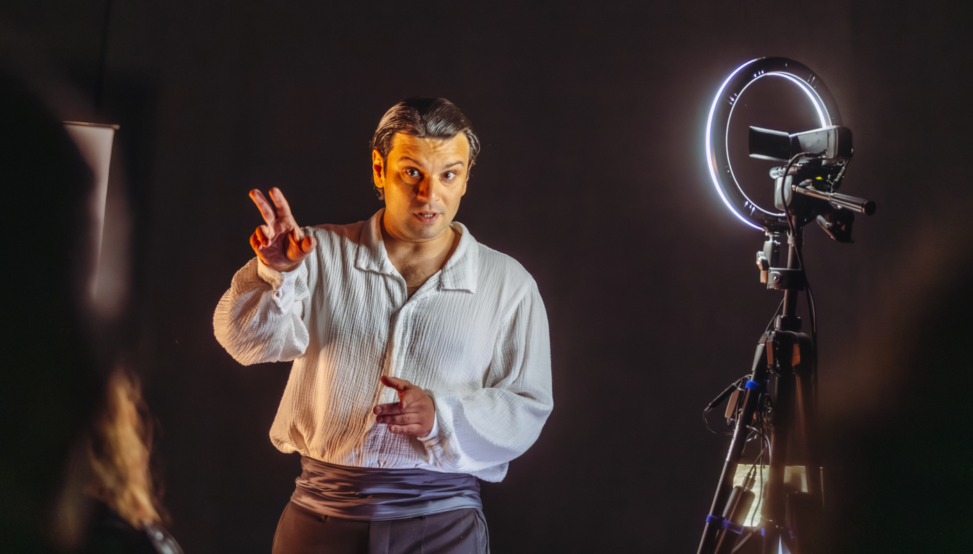 Zdjęcie mężczyzny w ujęciu od bioder w górę. Ma ciemne spodnie i jasną koszulę. Mężczyzna wyciąga przed siebie rękę, drugą trzyma blisko ciała na wysokości brzucha. Po prawej stronie stoi lampa na statywie, dająca blade światło. Tło czarne.