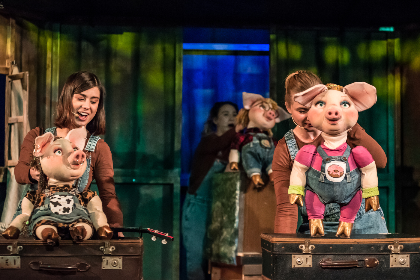 Zdjęcie aktorek animujących lalki w kształcie świnek w ubrankach. Po lewej świnka siedząca na dużej walizce, po prawej świnka stojąca na drugiej walizce, a za nią jeszcze dwie stojące w kolejce. Tło jest zielono-niebieskie.