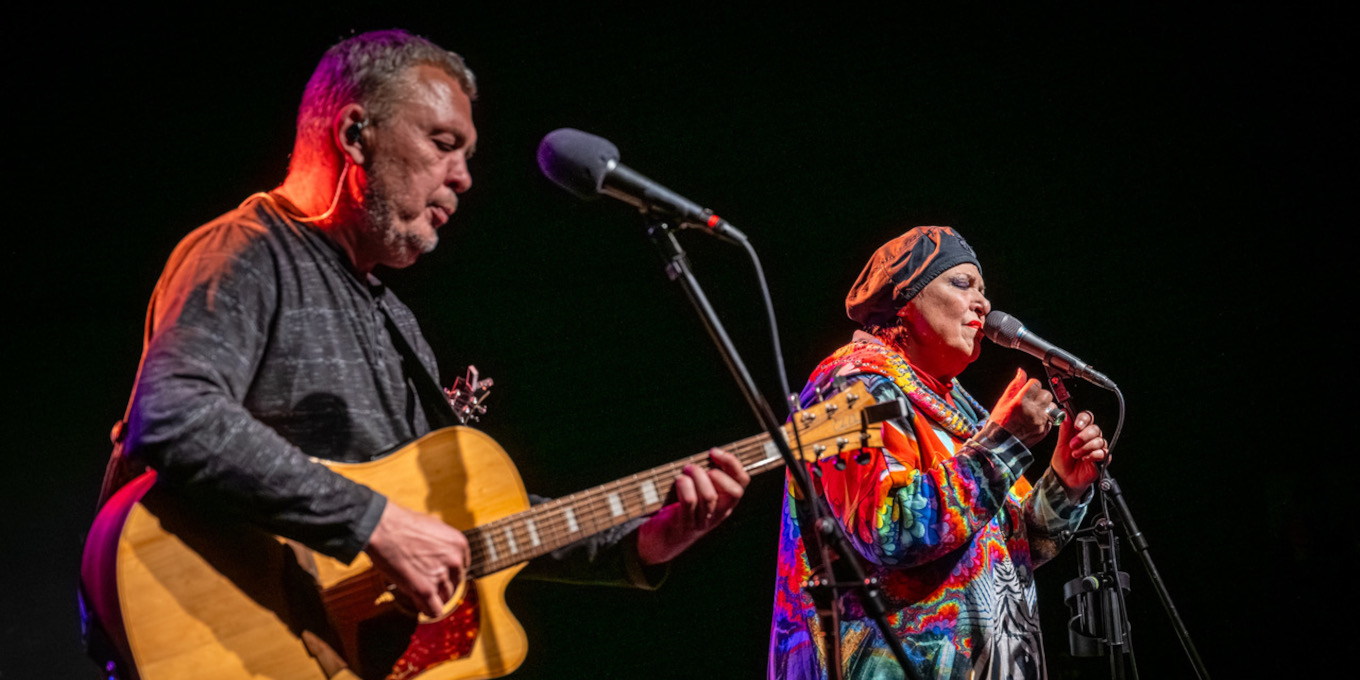 Dwie osoby na scenie. Po lewej mężczyzna z gitarą. Po prawej wokalistka w kolorowym stroju i berecie. Oboje mają przed sobą mikrofony na statywach.