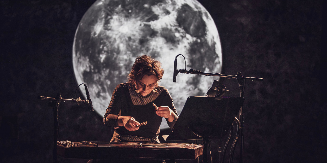 Zdjęcie kobiety grającej na cymbałach w pełnym skupieniu. Ma ciemny strój. Za nią jest projekcja z olbrzymim księżycem w pełni na ciemnym tle.
