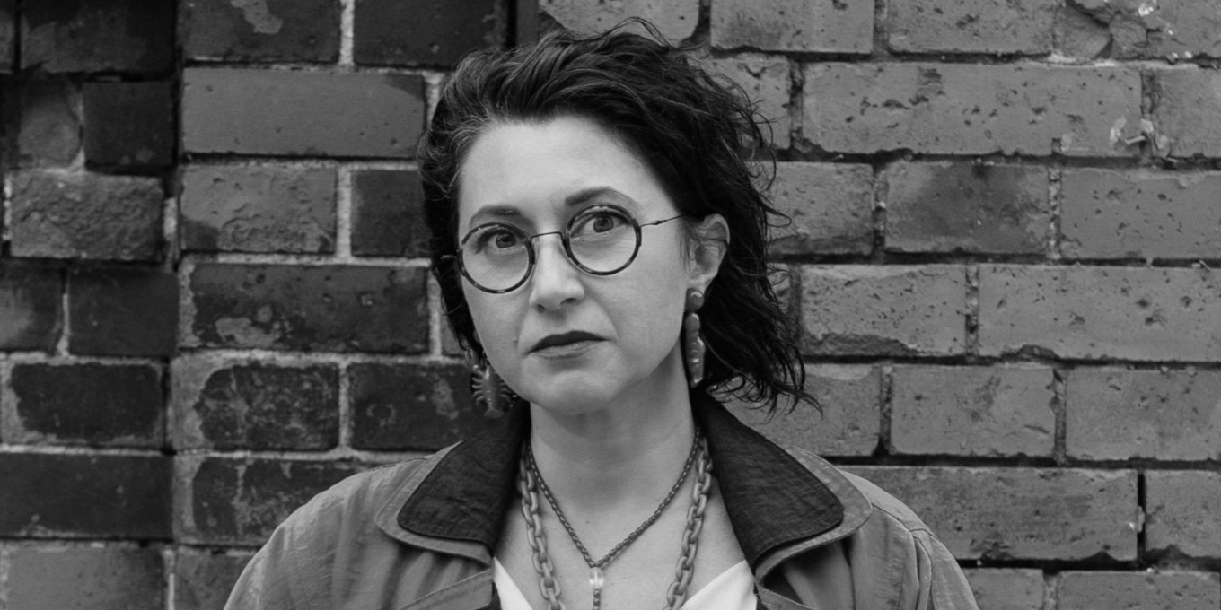 Czarno-białe zdjęcie zdjęcie kobiety w ujęciu portretowym. Kobieta ma ciemne włosy sięgające za ucho i okulary z okrągłymi oprawkami. W tle ściana z cegieł.